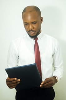 A man in a white shirt and red tie is holding and reading a document or folder. He appears to be focused and thoughtful with a neutral expression on his face. The background is plain white.