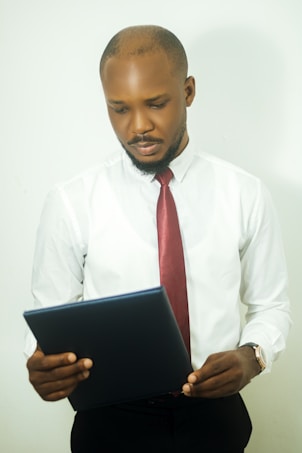 A man in a white shirt and red tie is holding and reading a document or folder. He appears to be focused and thoughtful with a neutral expression on his face. The background is plain white.