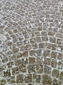 An arrangement of cobblestones forming an intricate circular pattern on a pathway. The stones have a weathered appearance with varying shades of gray and brown, and the gaps are filled with light-colored mortar.