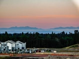 A scenic view featuring modern townhouse buildings in the foreground, surrounded by a construction site with visible equipment and materials. In the background, there's a dense forested area with mountains in the distance under a sky transitioning from light yellow to pink hues as the sun sets.