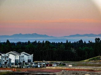 A scenic view featuring modern townhouse buildings in the foreground, surrounded by a construction site with visible equipment and materials. In the background, there's a dense forested area with mountains in the distance under a sky transitioning from light yellow to pink hues as the sun sets.