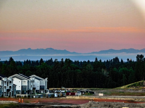 A scenic view featuring modern townhouse buildings in the foreground, surrounded by a construction site with visible equipment and materials. In the background, there's a dense forested area with mountains in the distance under a sky transitioning from light yellow to pink hues as the sun sets.
