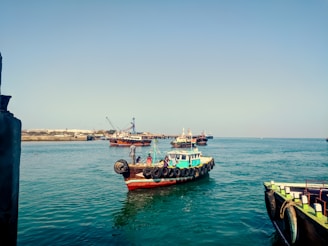 A small boat with tires along its sides floats on clear blue water. Several people can be seen on the boat. In the background, there are more boats, including larger vessels with cranes, near a shoreline with industrial structures. The sky is clear, and the water reflects its blue tint.