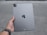 A person's hand is holding a silver Apple iPad. The back of the iPad is visible, showing the Apple logo, camera, and other design elements. The background appears to be an outdoor concrete or pavement surface.
