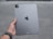 A person's hand is holding a silver Apple iPad. The back of the iPad is visible, showing the Apple logo, camera, and other design elements. The background appears to be an outdoor concrete or pavement surface.