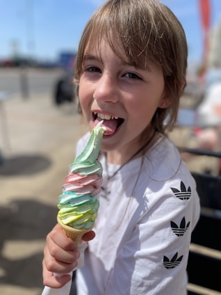 A young girl is happily licking a large, colorful swirl ice cream cone. She is wearing a white shirt with black logo designs on the sleeves. The background appears to be an outdoor setting, possibly a street or park, with blurry details of people and objects.