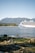 A large cruise ship with blue wave patterns is sailing across a calm body of water. In the background, there are lush green forests and tall mountains under a clear blue sky. Rocks and a patch of green mossy ground are visible in the foreground.