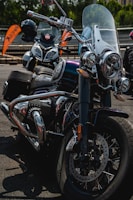 A close-up view of a classic style motorcycle with shiny chrome details and a large windshield. Behind it, another motorcycle is partially visible, parked in an outdoor setting with orange banners in the background.
