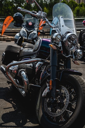 A close-up view of a classic style motorcycle with shiny chrome details and a large windshield. Behind it, another motorcycle is partially visible, parked in an outdoor setting with orange banners in the background.
