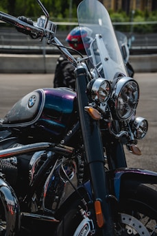 A close-up view of a motorcycle featuring a prominent chrome headlight, windshield, and a fuel tank with the BMW logo. The motorcycle is parked on a paved surface with some fencing and greenery visible in the background. The deep metallic colors of the motorcycle body include shades of blue and purple, with shiny chrome details.