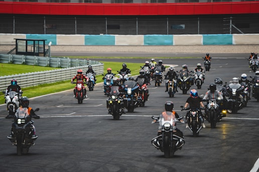 A large group of motorcyclists, wearing helmets and gear, rides together on a race track. The background features a railing, grass, and a section of red and white striped wall. The motorcyclists appear in formation, suggesting an organized event or rally.