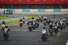 A large group of motorcyclists, wearing helmets and gear, rides together on a race track. The background features a railing, grass, and a section of red and white striped wall. The motorcyclists appear in formation, suggesting an organized event or rally.