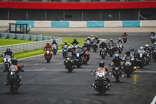 A large group of motorcyclists, wearing helmets and gear, rides together on a race track. The background features a railing, grass, and a section of red and white striped wall. The motorcyclists appear in formation, suggesting an organized event or rally.