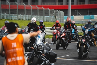 A group of motorcyclists wearing helmets and racing suits gather on a track, with a person in an orange vest directing them. The track is surrounded by metal barriers, and there is a grassy area in the background.