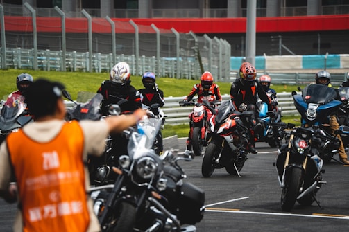 A group of motorcyclists wearing helmets and racing suits gather on a track, with a person in an orange vest directing them. The track is surrounded by metal barriers, and there is a grassy area in the background.