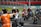 A group of motorcyclists wearing helmets and racing suits gather on a track, with a person in an orange vest directing them. The track is surrounded by metal barriers, and there is a grassy area in the background.