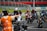 A group of motorcyclists wearing helmets and racing suits gather on a track, with a person in an orange vest directing them. The track is surrounded by metal barriers, and there is a grassy area in the background.