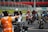 A group of motorcyclists wearing helmets and racing suits gather on a track, with a person in an orange vest directing them. The track is surrounded by metal barriers, and there is a grassy area in the background.