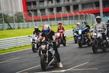 A group of motorcyclists wearing helmets and protective gear are lined up on a race track. The background features a building with red and gray accents and a series of barriers, likely for protection. The riders are in various positions, with one in the foreground appearing to be ready to accelerate.