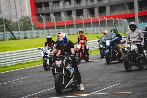 A group of motorcyclists wearing helmets and protective gear are lined up on a race track. The background features a building with red and gray accents and a series of barriers, likely for protection. The riders are in various positions, with one in the foreground appearing to be ready to accelerate.