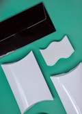 A flat lay composition of various packaging and design elements displayed on a green background. There is a black rectangular envelope, two white rectangular boxes with curved surfaces, and a white piece with wavy ridges. The items are positioned at diagonals, creating a dynamic layout.