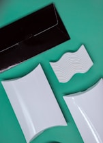 A flat lay composition of various packaging and design elements displayed on a green background. There is a black rectangular envelope, two white rectangular boxes with curved surfaces, and a white piece with wavy ridges. The items are positioned at diagonals, creating a dynamic layout.
