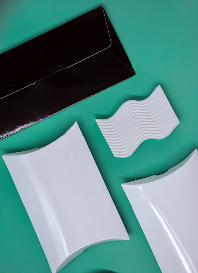 A flat lay composition of various packaging and design elements displayed on a green background. There is a black rectangular envelope, two white rectangular boxes with curved surfaces, and a white piece with wavy ridges. The items are positioned at diagonals, creating a dynamic layout.