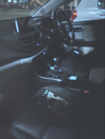 The interior of a car featuring a steering wheel, dashboard, and seats. A magazine or book is placed on the passenger seat. The lighting is dim, creating a low visibility atmosphere. The car appears to be parked indoors or in a garage.