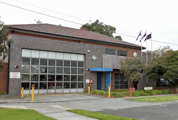 A two-story fire station building made of brick and glass, featuring large garage doors and a sign reading 'FIRE 000'. Three flags are raised on poles, and there are trees and a grassy area surrounding the building.