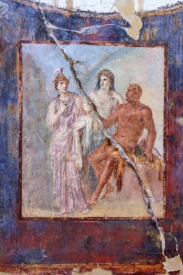 A fresco painting features three classical figures, two standing and one seated. The figures are elegantly dressed in flowing garments, with one figure wearing a helmet and holding a spear. The seated figure is partially nude, draped in a red cloth, with a muscular build. The background showcases faded colors and a cracked surface, indicating antiquity.