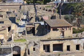 An archaeological site featuring well-preserved ancient structures with stone walls and tiled roofs. The area includes a mixture of open spaces and narrow pathways, with some trees and vegetation visible throughout the site. There are a few people walking and exploring the area.