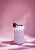 A creamy pink milkshake topped with whipped cream and a cherry, served in a glass with a straw against a matching pink background.