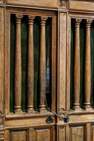 Wooden door with vertical columns and ornate carvings at the top. A chain and padlock are securing the door. The background features deep green fabric between the columns.