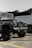 A rugged off-road vehicle is parked in an outdoor area with a raised hood and large tires. The vehicle's exterior is a dark color, possibly green or grey, with a prominent front grille and round headlights. In the background, there is a building with white walls and large windows, and some foliage is visible.