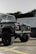 A rugged off-road vehicle is parked in an outdoor area with a raised hood and large tires. The vehicle's exterior is a dark color, possibly green or grey, with a prominent front grille and round headlights. In the background, there is a building with white walls and large windows, and some foliage is visible.