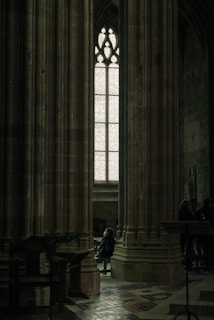 A person sits alone in a dimly lit, grand cathedral with towering stone columns and an ornate stained-glass window. The light filtering through casts a serene atmosphere, creating a sense of solitude and introspection.