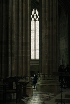 A person sits alone in a dimly lit, grand cathedral with towering stone columns and an ornate stained-glass window. The light filtering through casts a serene atmosphere, creating a sense of solitude and introspection.