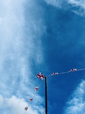A string of Union Jack flags is strung across a tall pole against a backdrop of a vivid blue sky with scattered clouds.