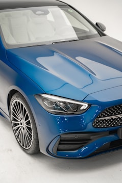 A close-up view of the front section of a sleek, modern car with a metallic blue finish. The focus is on the headlight, grille, and front tire, showcasing the vehicle's stylish design and high-quality craftsmanship.