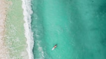 Aerial view of a person paddleboarding across clear turquoise waters near a sandy shoreline. White waves gently crash against the shore, creating a tranquil and expansive scene.