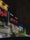 Several storefronts with brightly lit signs in Arabic are seen. The signs are colorful, featuring red, blue, and yellow letters. The facades have multiple lights and air conditioning units attached. The ambient lighting suggests a nighttime setting, giving the scene an urban look.