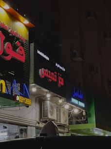 Several storefronts with brightly lit signs in Arabic are seen. The signs are colorful, featuring red, blue, and yellow letters. The facades have multiple lights and air conditioning units attached. The ambient lighting suggests a nighttime setting, giving the scene an urban look.