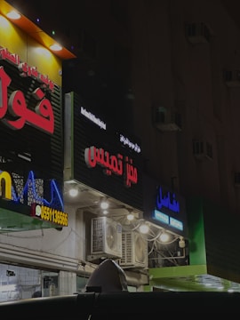Several storefronts with brightly lit signs in Arabic are seen. The signs are colorful, featuring red, blue, and yellow letters. The facades have multiple lights and air conditioning units attached. The ambient lighting suggests a nighttime setting, giving the scene an urban look.