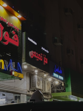 Several storefronts with brightly lit signs in Arabic are seen. The signs are colorful, featuring red, blue, and yellow letters. The facades have multiple lights and air conditioning units attached. The ambient lighting suggests a nighttime setting, giving the scene an urban look.