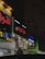 Several storefronts with brightly lit signs in Arabic are seen. The signs are colorful, featuring red, blue, and yellow letters. The facades have multiple lights and air conditioning units attached. The ambient lighting suggests a nighttime setting, giving the scene an urban look.
