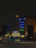 A city street at night with neon signs in Arabic and English on a building. Several cars are parked and moving on the road, and a streetlight casts a warm glow.
