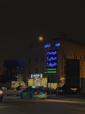 A city street at night with neon signs in Arabic and English on a building. Several cars are parked and moving on the road, and a streetlight casts a warm glow.