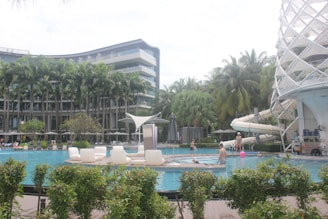 A luxurious swimming pool area surrounded by tall palm trees and a modern multi-story hotel. Several people are relaxing and playing in the pool, with some sitting on poolside loungers. A water slide structure is visible on the right side, and there are sun umbrellas and various greenery enhancing the tropical setting.