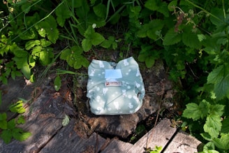 A green diaper with a pattern featuring white bunnies and other small shapes is placed on a tree stump in a natural setting. The surroundings are filled with lush green leaves and grass, with some wooden planks visible on the ground.