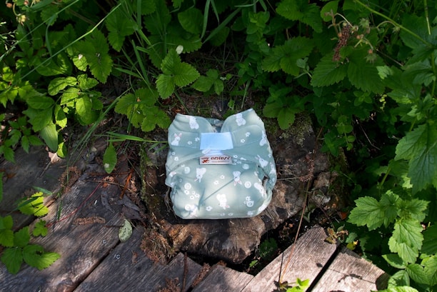A green diaper with a pattern featuring white bunnies and other small shapes is placed on a tree stump in a natural setting. The surroundings are filled with lush green leaves and grass, with some wooden planks visible on the ground.